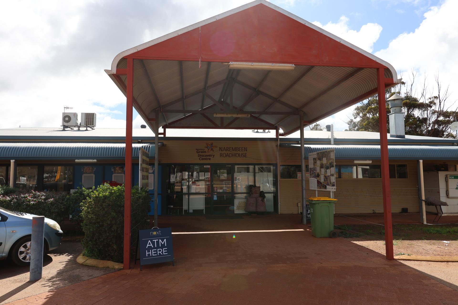 A building entrance with a red roof, featuring signs for an ATM and a grain discovery center. A blue car is parked nearby.